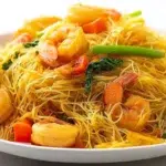 Singapore noodles stir-fried with rice noodles, vegetables, shrimp, and curry spices in a colorful wok dish.
