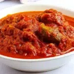 A bowl of rich, deep-red Nigerian tomato stew made with tomatoes, peppers, and onions, served alongside rice.