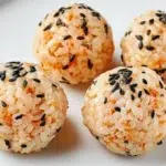 Four no-cook salmon rice balls coated in black and white sesame seeds, arranged on a white plate.