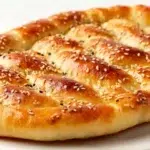 Golden-brown Turkish pide flatbread with a braided crust, topped with sesame and nigella seeds, on a white plate.
